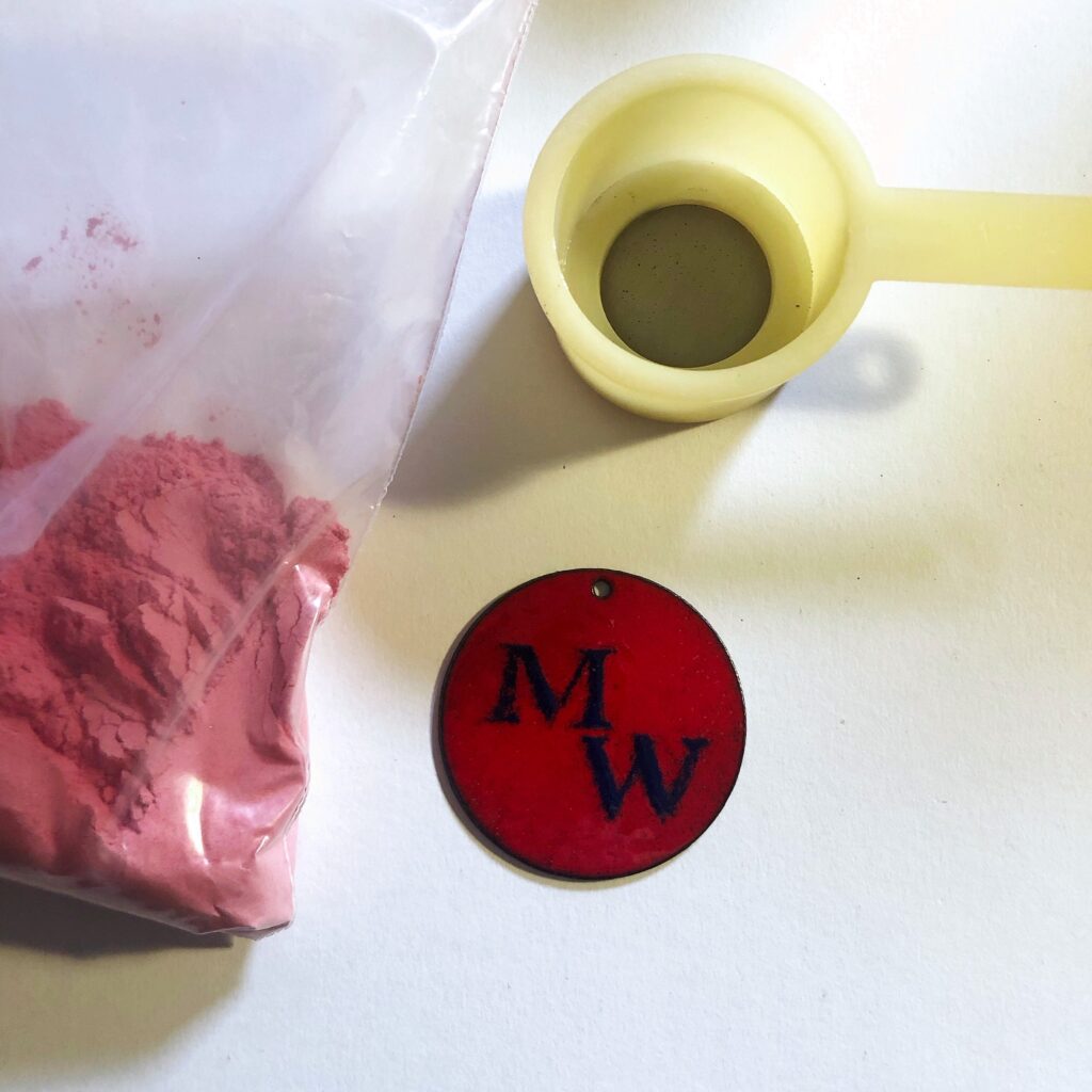 Ein runder, roter Emaille-Anhänger mit den dunkelblauen Initialen MW, daneben eine Plastiktüte mit rotem Emaillepulver und ein kleines Sieb auf weißem Untergrund.